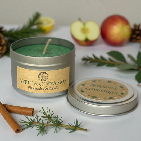 Handmade soy candle in a tin with apple and cinnamon theme, surrounded by apples, cinnamon sticks, and greenery.